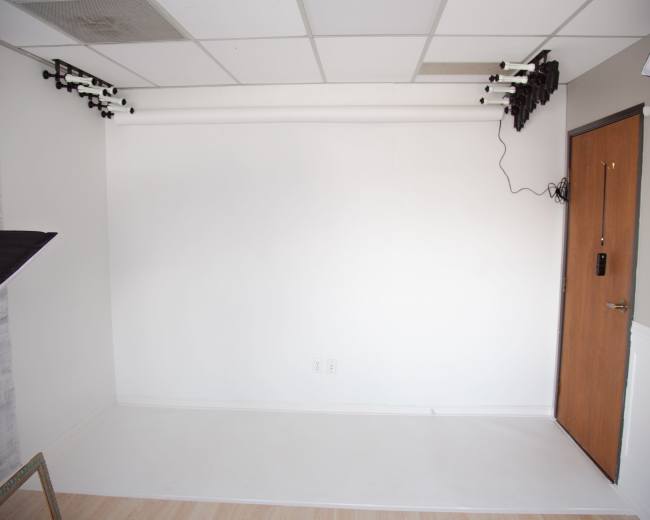The image shows a minimalistic studio space with a white backdrop, a door on the right, and lighting equipment mounted on the ceiling.