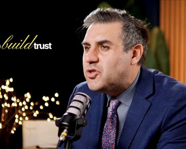 A man in a suit is speaking into a microphone in a studio setting with soft lighting and decorative plants in the background.