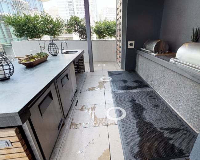 The image shows a modern outdoor kitchen area with a sink, grill, and tiled flooring surrounded by city buildings.