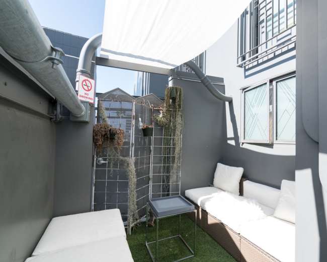 A small outdoor seating area features white cushions on two benches, a metal lattice with hanging plants, and a table under a rooftop covering.