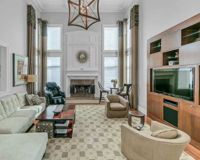 The living room features a sofa, recliner, and armchair arranged around a coffee table in front of a fireplace, with large windows allowing natural light and a television mounted on a wooden panel.