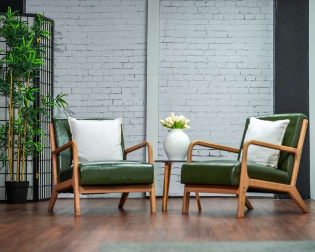 The image shows two green armchairs with wooden frames facing each other, accompanied by a small round table with a white vase holding flowers, set against a backdrop of a brick wall and a decorative plant.