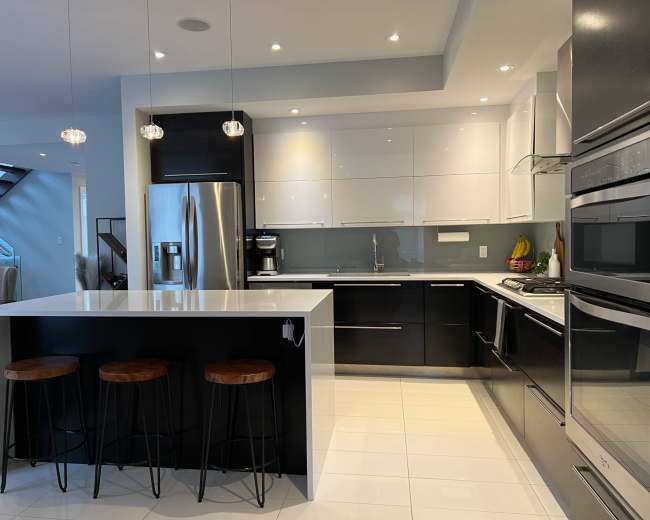 The image shows a modern kitchen featuring sleek black and white cabinetry, stainless steel appliances, and three bar stools at a white island counter.