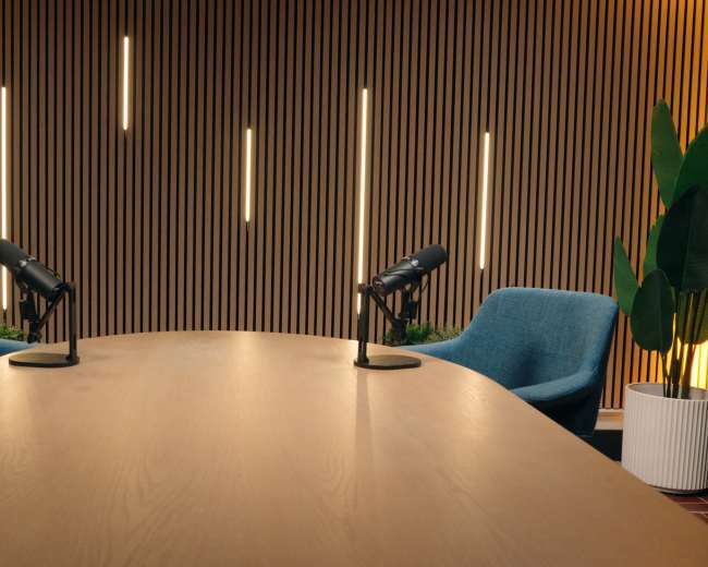 A large wooden table sits between two blue chairs, each equipped with a microphone, set against a wall with vertical wooden slats and decorative lighting.