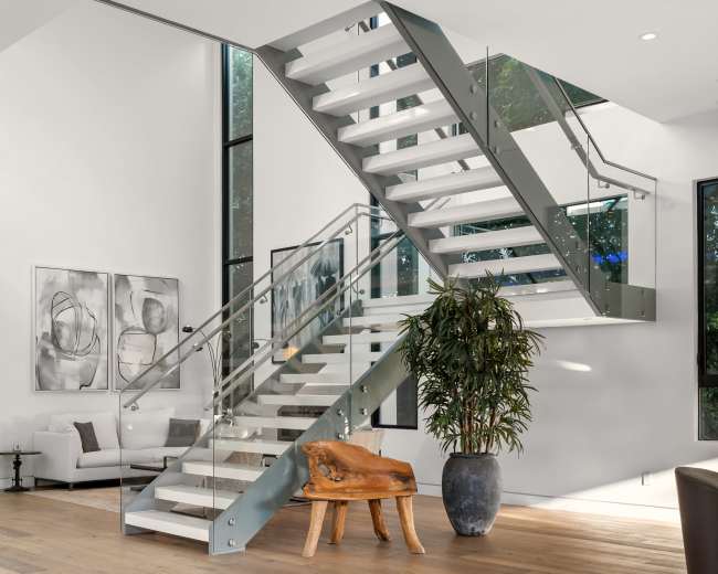 The image shows a modern interior featuring a glass and metal staircase, surrounded by large windows and minimalist furniture.