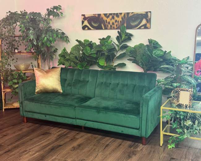 The image shows a green velvet sofa with a gold accent pillow, positioned among various indoor plants and decorated with a gold side table.