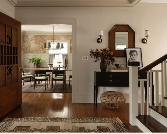 The image shows a well-lit entryway with a wooden front door, leading to a dining area visible through an open doorway, featuring a table and chairs.