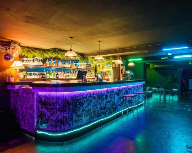 A brightly lit bar featuring colorful neon lights, shelves stocked with various bottles, and a tropical-themed decor.