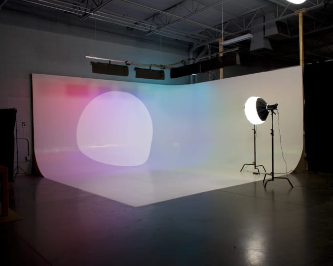 The image shows a sparse photo studio with a gradient backdrop and a large light source positioned on a stand.