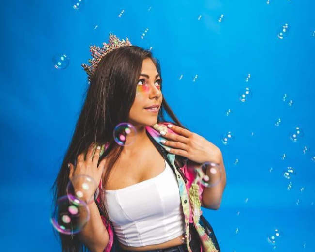 A person wearing a crown and colorful clothing sits crossed-legged against a blue backdrop, surrounded by floating bubbles.