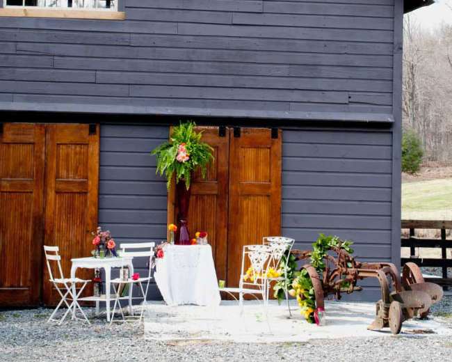 A rustic barn features wooden double doors, a small table adorned with flowers, and white chairs set up outdoors.