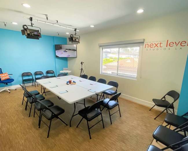 The image shows a brightly lit conference room with a large table surrounded by black chairs, featuring a television mounted on the wall and a blue accent wall.
