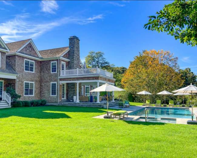 The image shows a spacious, two-story house with a swimming pool and several lounge chairs on a well-manicured lawn.