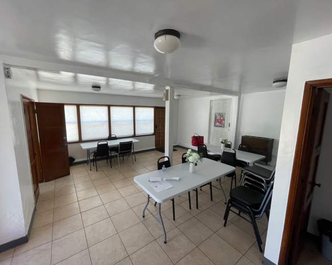 The image shows a bright, empty room furnished with several tables and chairs arranged in a meeting setup, with large windows allowing natural light to enter.
