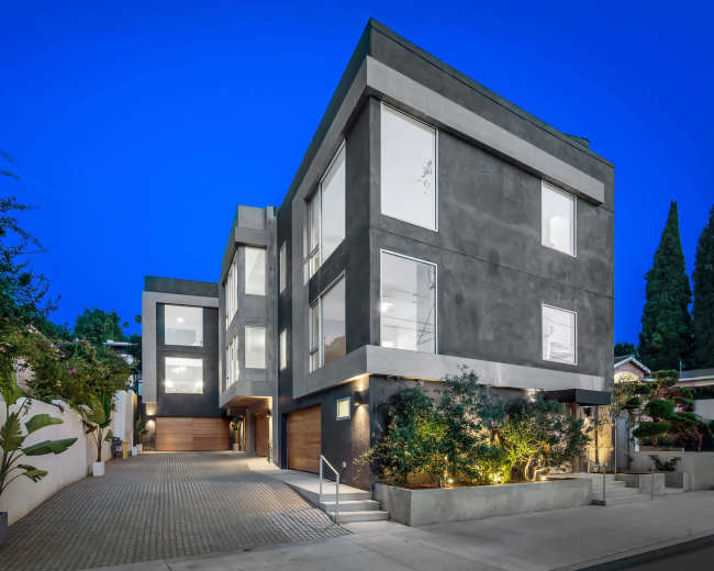 The image shows a modern, multi-story house with large windows, situated on a tree-lined street at dusk.