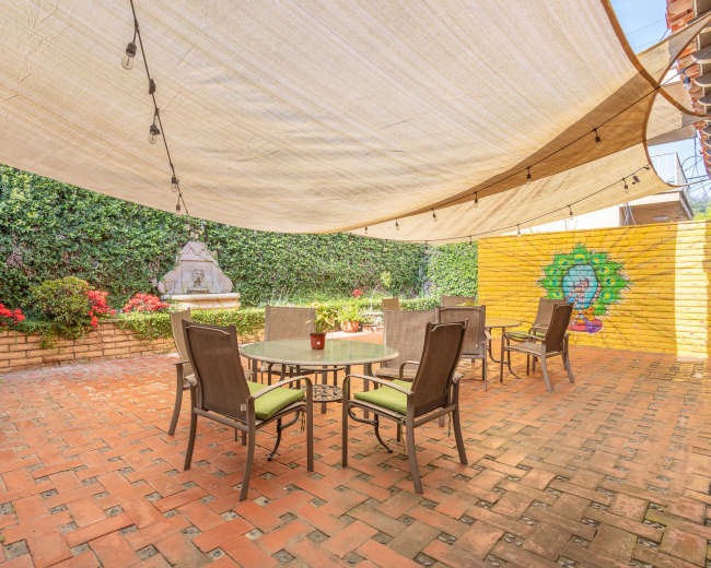 A patio area is shaded by a large canopy and features a round table with six chairs, surrounded by vibrant flowers and a mural on the wall.