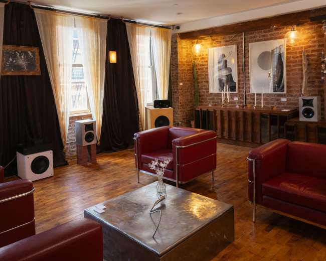 The image shows a cozy art gallery with red leather chairs, wooden flooring, and several framed artworks displayed on the brick walls.