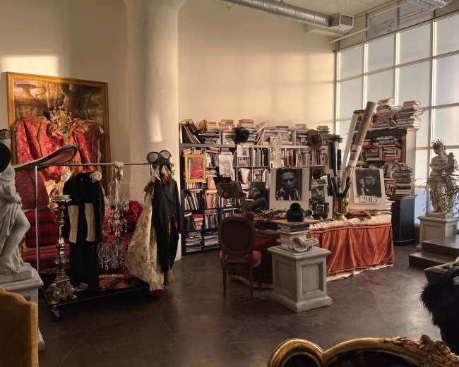 A spacious room filled with bookshelves, clothing on display, and various artistic decorations, including a large painting and sculptures.