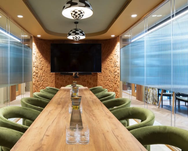 A modern conference room features a long wooden table surrounded by green chairs, with decorative walls and a large screen at one end.