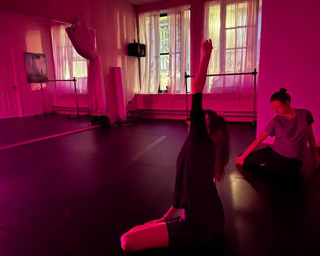 Two dancers practice in a brightly lit studio with pink-tinted lighting, one sitting with an arm raised and the other stretching nearby.
