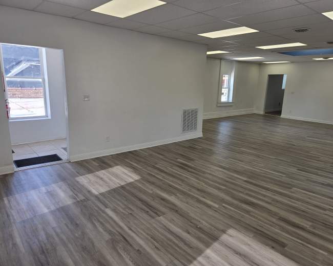 The image shows a spacious, empty room with light hardwood flooring, white walls, and large windows.
