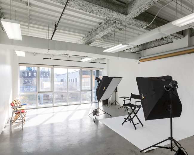 The image shows a photography studio equipped with two lighting setups, a backdrop, and a director's chair near large windows that allow natural light.