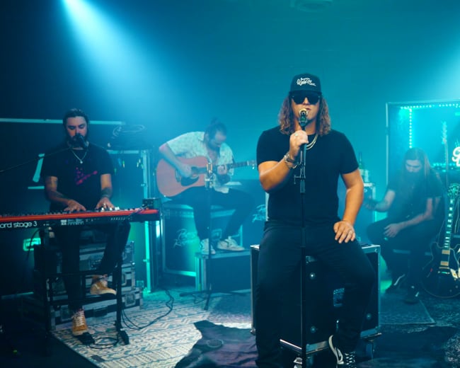 A musician sits in front of a microphone while a band plays acoustic instruments in a dimly lit setting with colorful lights.