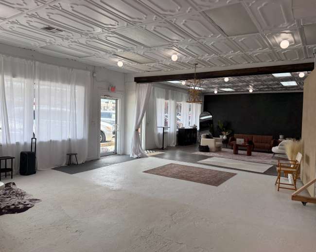 The image shows a spacious, well-lit interior with white walls, a sectional couch, and various furniture pieces arranged in an open layout.