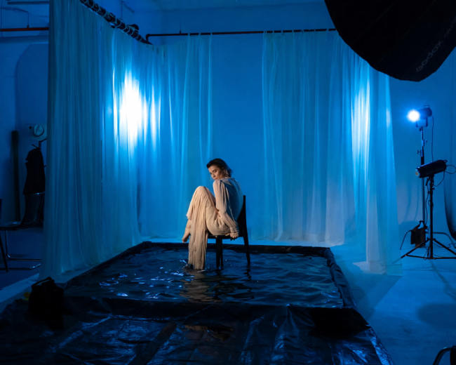 A model sits on a chair in a partially flooded studio surrounded by blue lighting and draped fabric.