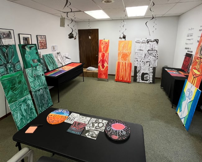 The room features several large and colorful paintings displayed on easels and tables, with black and white streamers hanging from the ceiling.