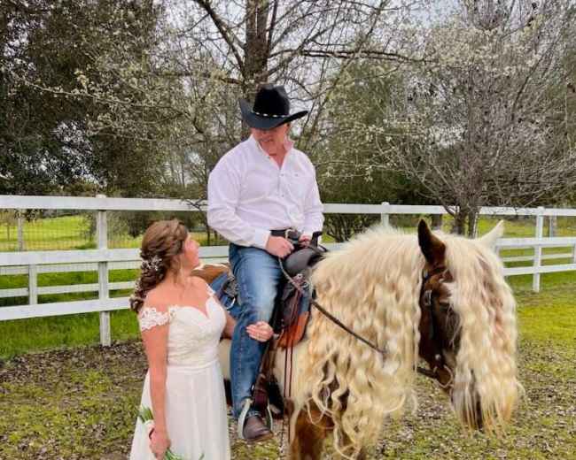 A woman in a white wedding dress stands beside a man in a cowboy hat seated on a horse with long, flowing hair.