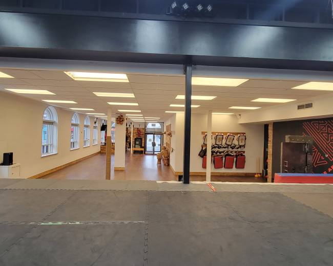 The image shows the interior of a spacious martial arts studio with an open area, large windows, and workout equipment along the walls.