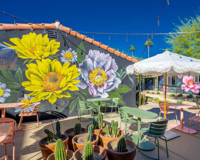 The image features a vibrant outdoor seating area with colorful chairs and tables surrounded by large murals of flowers and potted cacti under a white canopy.