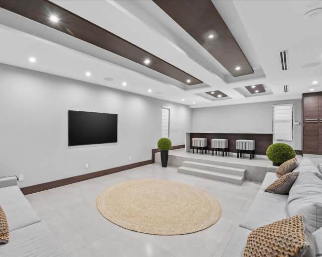 A modern living room features a large area rug, grey sofas, a mounted television, and a bar-style seating area with high chairs.