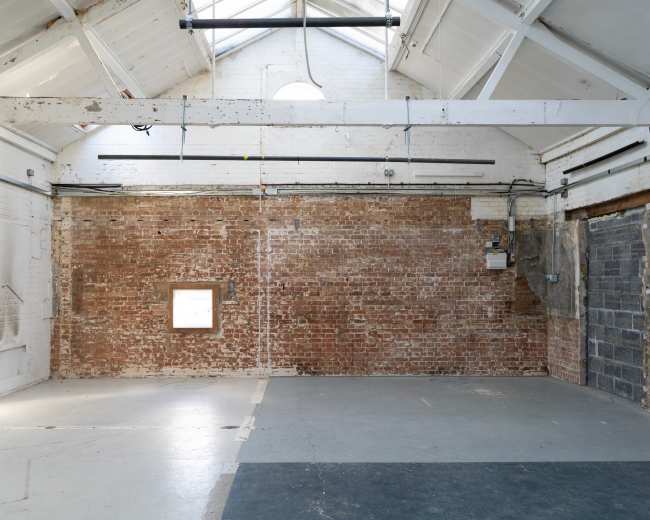 The image shows an empty warehouse interior with exposed brick walls, a high ceiling, and a concrete floor.