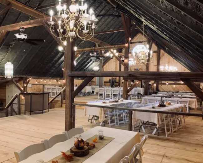 The image shows a spacious, rustic barn interior with wooden beams, a chandelier, and banquet tables set for an event.