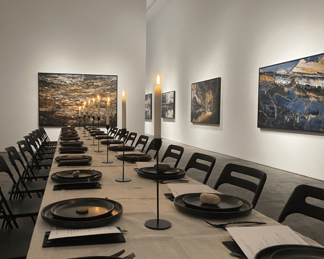A long, empty dining table is set in a gallery surrounded by various paintings on the walls.
