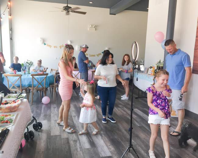 A group of people is gathered in a room decorated for a celebration, with tables set for food and balloons, as children play and adults mingle.