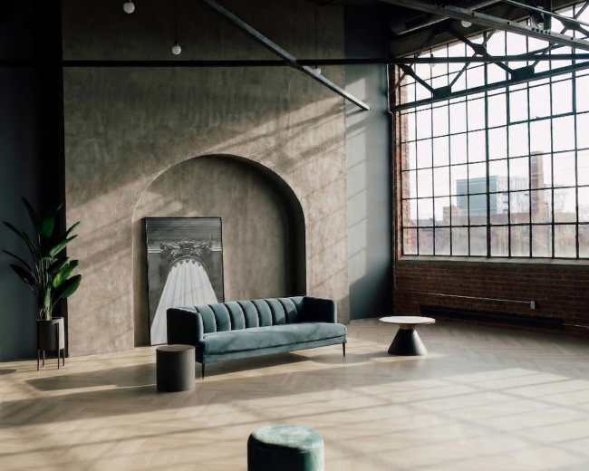 The image features a spacious, modern interior with a velvet sofa, a plant, circular side tables, and large windows allowing natural light to flood the room.