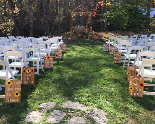 A pathway lined with white chairs and wooden signs featuring inspirational quotes leads to a decorated altar in a forested outdoor setting.