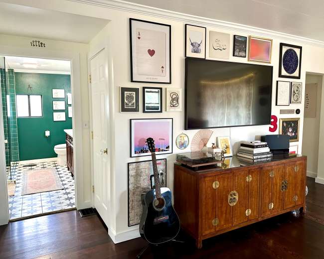 The image shows a living area featuring a wooden cabinet adorned with books and a guitar, alongside a gallery wall of framed art, with an open doorway leading to a bathroom visible in the background.