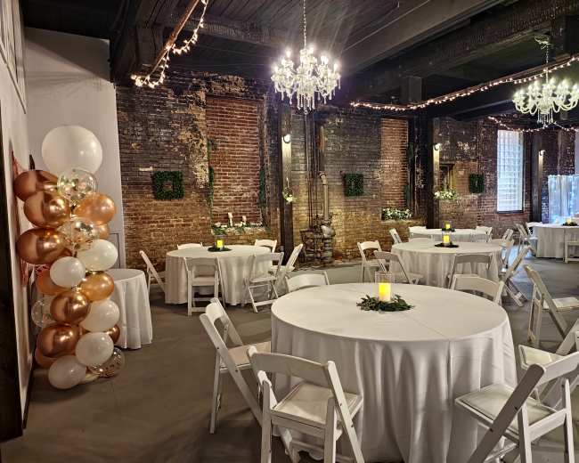 A decorated event space with round tables covered in white tablecloths, chandeliers hanging from the ceiling, and a festive balloon arrangement in one corner.