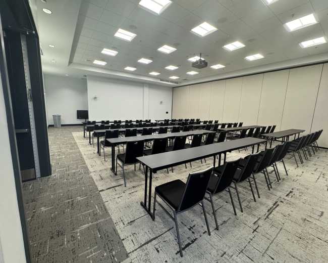 A conference room features rows of black tables and chairs arranged in a grid pattern, with light-colored walls and a projector mounted on the ceiling.