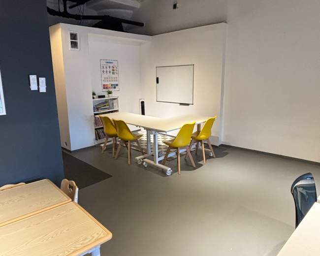 The image shows a modern, minimalistic room with a table and four yellow chairs, a whiteboard on the wall, and light-colored furniture.