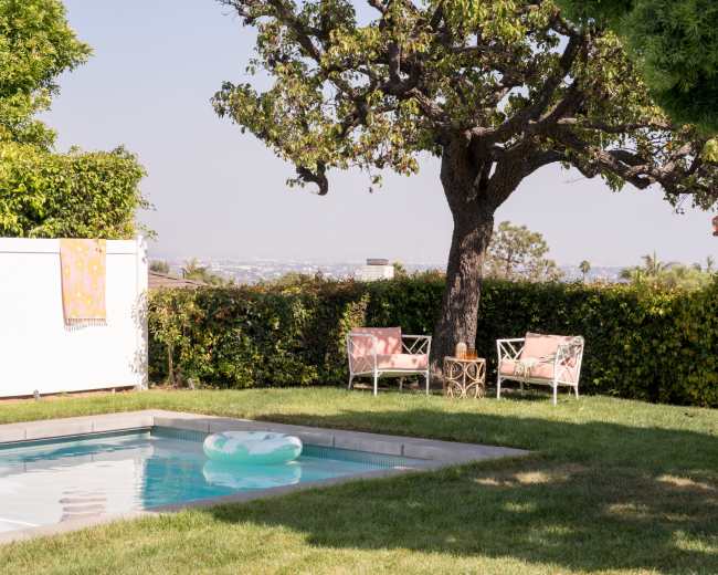 A clear swimming pool is surrounded by a grassy area and a large tree, with two chairs placed nearby and a colorful towel hanging on the fence.