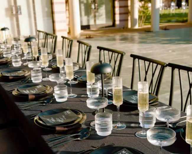 A long dining table is set outdoors with elegant black plates, clear glassware, and cutlery, surrounded by greenery.