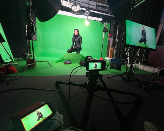 A person sits on a chair in front of a green screen, surrounded by studio lighting and equipment.