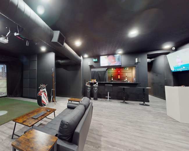 The image shows an indoor golf simulation room featuring a large projection screen, a putting green, a bar area, and seating for spectators.