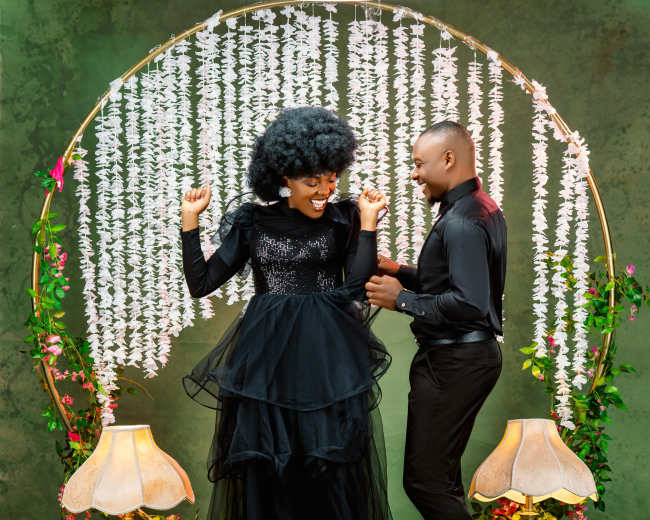 A man and woman dance joyfully in front of a decorative floral backdrop, illuminated by two vintage lamps.