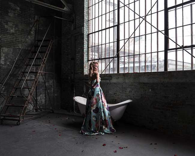 A woman in a floral gown stands beside a vintage bathtub in a large, industrial room with tall windows and a metal ladder.
